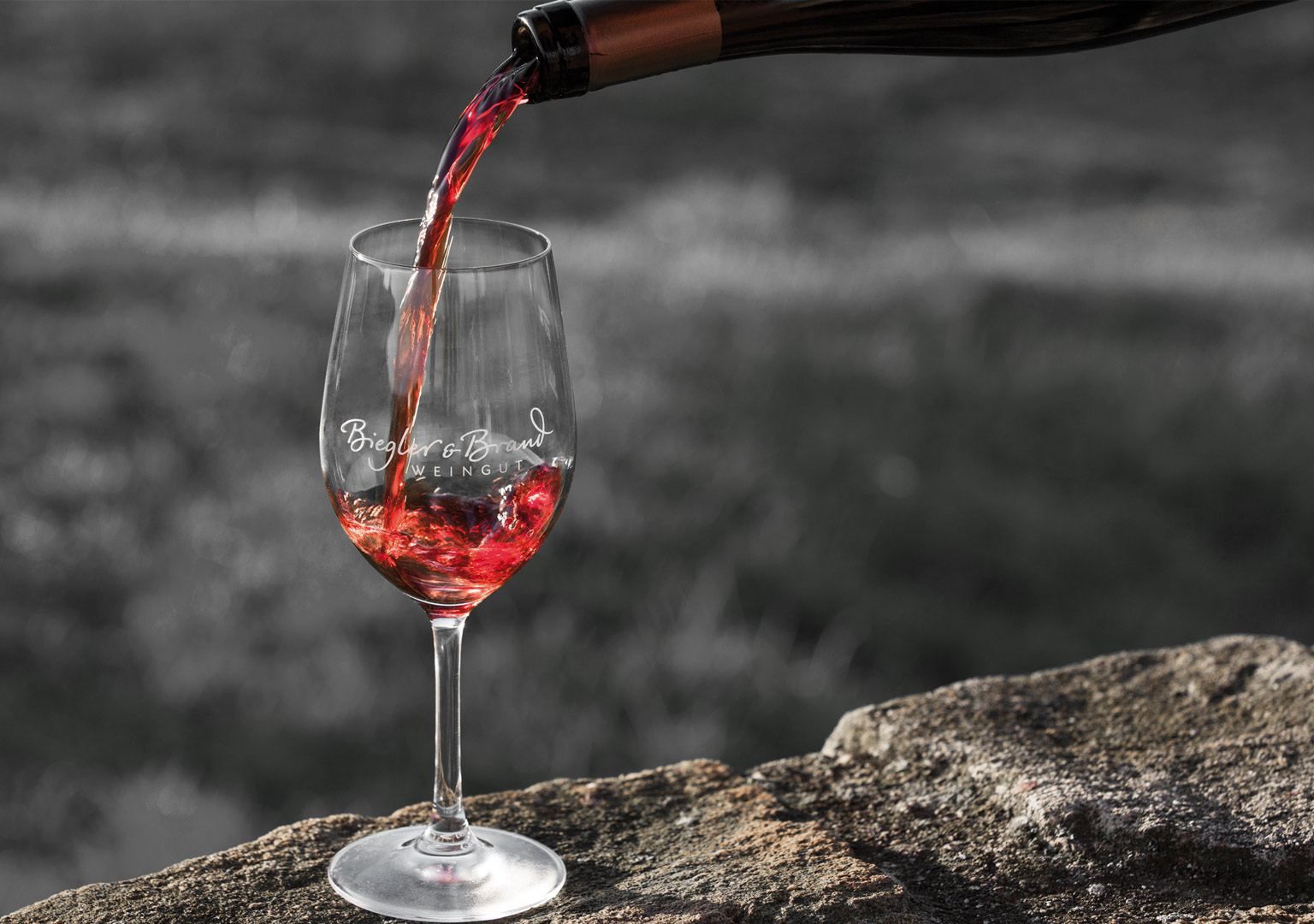 Ein Glas mit Logo vom Weingut Biegler & Brand steht in den Weinbergen auf einem Stein auf einem in das Glas wird Rotwein eingeschenkt 